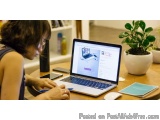 The image shows a young woman sitting at a wooden table with a laptop in front of her. She is holding a pen and appears to be...