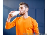 The image shows a young man drinking milk from a bottle. He is wearing an orange long-sleeved shirt and has a beard. The back...