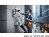 The image shows a man on a ladder installing a security camera on the side of a building. He is wearing a blue shirt and a to...
