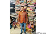 The image shows a young man standing in front of a large display of colorful fabrics. He is wearing a traditional Indian outf...