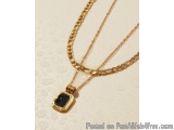 The image shows a gold necklace with a pendant. The pendant is square-shaped and has a black stone in the center. The necklac...