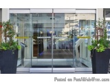 The image shows the entrance of a modern building with a large glass door. The door is made of stainless steel and has a yell...