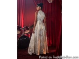The image shows a woman standing in front of a red curtain with a drum set in the background. She is wearing a long, flowing,...