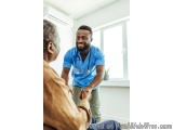 The image shows a young African-American male doctor wearing a blue scrub suit and a stethoscope around his neck, standing in...