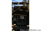 The image shows a two-story house with a black exterior. The house has a sloping roof and a small porch with a few chairs and...
