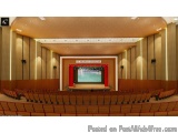 The image shows a large auditorium with a high ceiling and wooden walls. The auditorium has rows of red seats facing the stag...