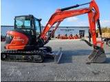 The image shows a red Kubota KX0575 mini excavator parked in an empty parking lot. The excavator has a long arm with a bucket...