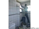 The image shows two men working on a tiled wall in a bathroom. They are standing on a ladder and appear to be in the process...