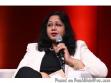 The image shows a woman sitting on a white couch and speaking into a microphone. She is wearing a white blazer and a black to...