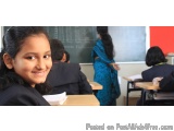 The image shows a young girl sitting at a desk in a classroom. She is wearing a black blazer and has a smile on her face. Beh...
