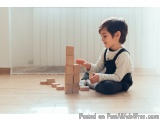 The image shows a young boy sitting on the floor in a room with white walls and wooden flooring. He is wearing a white shirt...