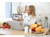 The image shows a young woman in a white lab coat sitting at a kitchen table with a laptop and a plate of fresh fruits and ve...