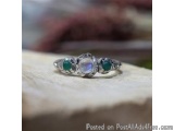 The image is a close-up of a ring on a piece of wood. The ring is made of silver and has three stones on it. The center stone...
