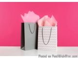The image shows two gift bags on a pink background. The bag on the left is grey and the one on the right is white with a chev...