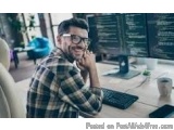 The image shows a man sitting at a desk in front of a computer. He is wearing a plaid shirt and glasses and has a beard. He h...