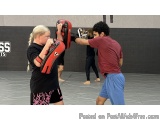 The image shows a man and a woman in a martial arts class. The woman is wearing a black t-shirt and pink shorts with a red an...