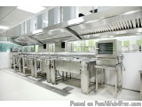 The image shows a modern commercial kitchen with a long row of stainless steel sinks and a large range hood above them. The k...