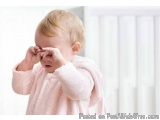 The image shows a baby girl standing in front of a white radiator. She is wearing a pink fluffy robe and has blonde hair. The...
