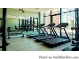 The image shows a modern and well-equipped gym with a row of treadmills and elliptical machines. The gym has large windows th...
