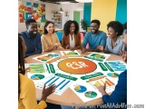 The image shows a group of seven young people sitting around a round table in a meeting room. They are all smiling and appear...