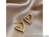 The image shows a pair of gold earrings lying on a beige satin fabric. The earrings are in the shape of a heart, with a curve...