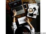 The image shows a person sitting at a wooden table with a laptop, a notebook, a cup of coffee, a magazine, and a pair of head...