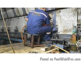 The image shows a man working on a large industrial machine in a workshop. He is wearing a blue coverall with a yellow logo o...