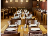 The image shows a long wooden dining table set up in a restaurant. The table is set with white plates, wine glasses, and cutl...
