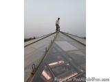 The image shows a man standing on the edge of a glass roof of a building. He is wearing a white t-shirt, blue jeans, and a bl...