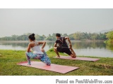The image shows a man and a woman performing yoga on pink yoga mats on a grassy area near a lake. The woman is wearing a whit...