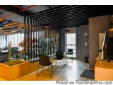 The image shows a modern office space with a high ceiling and a large window on the right side. The window has a black and wh...