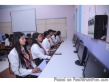 The image shows a group of young students sitting at desks in a computer lab. They are all wearing white lab coats and are fo...
