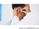 The image shows a close-up of a man's ear. He is holding a hearing aid in his right hand and adjusting it with his left hand....