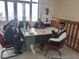 The image shows a group of six men sitting around a conference table in a room with large windows. They are all dressed in fo...