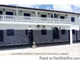 The image shows a two-story white building with a black fence in front of it. The building has a balcony on the second floor...