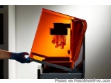 The image shows a person's hand wearing a blue glove holding a 3D printer. The printer is orange in color and has a sleek, mo...