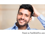 The image is a close-up portrait of a young man with a big smile on his face. He is wearing a light blue collared shirt and h...