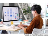 The image shows a young man sitting at a desk in front of a computer monitor. He is wearing a brown shirt and appears to be w...