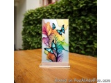 The image is a photograph of a small acrylic stand on a wooden table. The stand is rectangular in shape and has a white base....