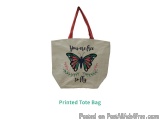 The image is of a printed tote bag. The bag is light beige in color and has a red handle. On the front of the bag, there is a...