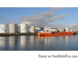 The image shows a large orange cargo ship docked at a port. The ship is in the center of the image, with a large white tanker...