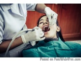 The image shows a young girl sitting in a dental chair with her mouth open. She is wearing a green dental gown and is being e...