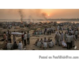 The image shows a large group of people gathered on a beach at sunset. The sky is orange and the sun is setting in the backgr...