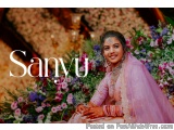 The image is a portrait of a young Indian woman, Sanyu, sitting on a chair in front of a floral backdrop. She is wearing a tr...