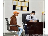 The image shows a young man sitting at a desk in an office. He is wearing a white lab coat and a face mask, and is holding a...