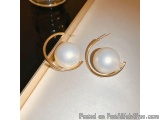 The image shows a pair of gold earrings with a white pearl in the center. The earrings are circular in shape and have a hook...