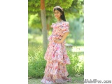 The image shows a young girl standing in a garden. She is wearing a long, flowy dress with a pink floral print. The dress has...