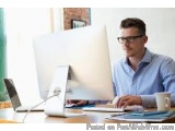 The image shows a young man sitting at a desk in front of a computer monitor. He is wearing a blue shirt and glasses and appe...