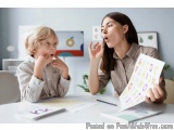 The image shows a mother and son sitting at a desk in an office. The mother is holding a piece of paper with colorful illustr...