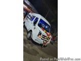 The image shows a white van parked on a street at night. The van is decorated with a garland of red and yellow flowers and ga...